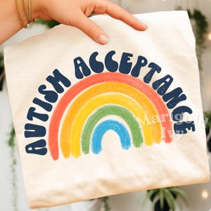 May include: Cream-colored t-shirt featuring the words "AUTISM ACCEPTANCE" in navy blue, arched above a rainbow design. The rainbow includes bands of red, orange, yellow, green, and blue. The shirt is held by a person.