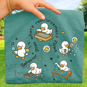 May include: Teal-colored fabric featuring a cartoon duck design. The design includes ducks playing with bubbles, maracas, and a pop-it toy. Text around the ducks reads "where reinforcement goes behavior flows."