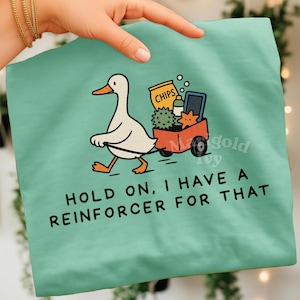 May include: A light teal t-shirt features a cartoon duck pulling a red wagon filled with snacks. The wagon contains a bag of chips, a bottle, a star, and a spiky ball. The text "HOLD ON, I HAVE A REINFORCER FOR THAT" is printed below.