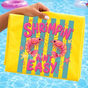 May include: Yellow beach towel with the text "Shrimpin' Ain't Easy" in pink, featuring two pink shrimp and blue vertical stripes. The towel also has pink starfish and yellow accents.