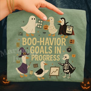 May include: A light green t-shirt featuring cartoon ducks dressed in Halloween costumes. The shirt has the text "BOO-HAVIOR GOALS IN PROGRESS". The ducks are dressed as a ghost, mummy, penguin, witch, and skeleton.