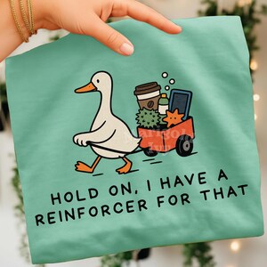 May include: A light green t-shirt featuring a graphic of a white duck pulling a red wagon filled with a coffee cup, a green bottle, a spiky green ball, and an orange starfish. The text reads: HOLD ON, I HAVE A REINFORCER FOR THAT.