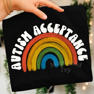 May include: Black t-shirt with a rainbow design and the words "AUTISM ACCEPTANCE" in white, arched above the rainbow. The rainbow features red, orange, yellow, green, and blue arcs. The shirt is held by a hand.
