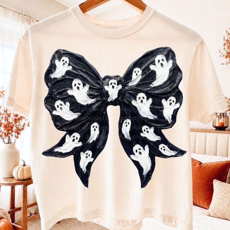 K&ouml;nnte beinhalten: Ein cremefarbenes Kurzarm-T-Shirt mit einer gro&szlig;en, mittigen Grafik einer schwarzen Schleife. Die Schleife ist mit zahlreichen kleinen, wei&szlig;en, handgemalt wirkenden Geistern mit ausdrucksstarken Gesichtern auf der dunklen Textur verziert.