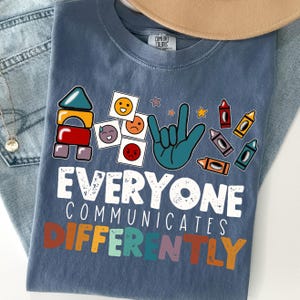 May include: Blue Comfort Colors t-shirt with a graphic design. The design includes building blocks, emoji faces, a hand in sign language, and crayons. The text reads "EVERYONE COMMUNICATES DIFFERENTLY."