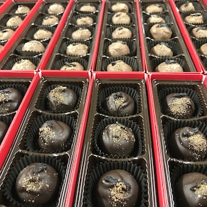 May include: An overhead shot of several red boxes filled with chocolate truffles. The truffles are dark brown and round, some dusted with a light-colored powder. Each truffle sits in a fluted paper cup within the box.