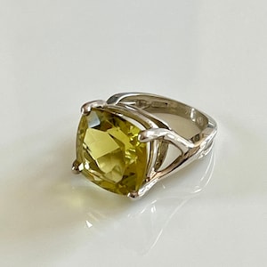 May include: A silver ring featuring a large, square-cut, light green gemstone. The ring has a unique, openwork design on the band, with the gemstone held in place by four prongs. The ring is set against a white background.