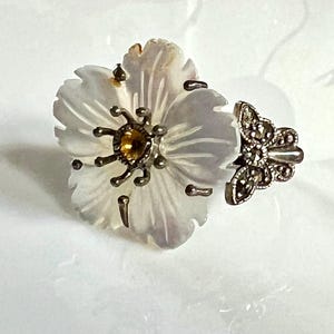 May include: A vintage flower-shaped ring with a white mother-of-pearl petal design. The center features a yellow gemstone, and the ring has a silver-toned metal setting. The ring's band has a decorative butterfly design.