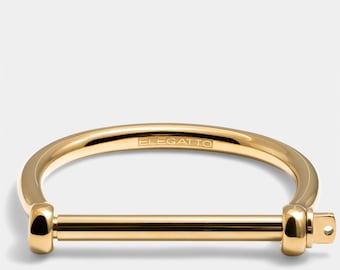 May include: A gold-toned metal bracelet with a simple, minimalist design. The bracelet features a thick, rounded band with a straight bar that acts as a clasp. The inscription "ELEGATTO" is visible on the inside of the band.
