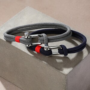 May include: Two braided rope bracelets with silver clasps. The top bracelet is gray with a red accent and the bottom bracelet is navy blue.