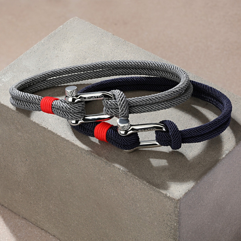 May include: Two braided rope bracelets with silver clasps. The top bracelet is gray with a red accent and the bottom bracelet is navy blue.