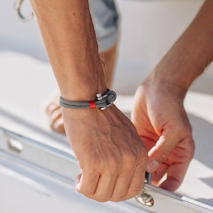May include: A gray rope bracelet with a red accent and a silver anchor clasp.