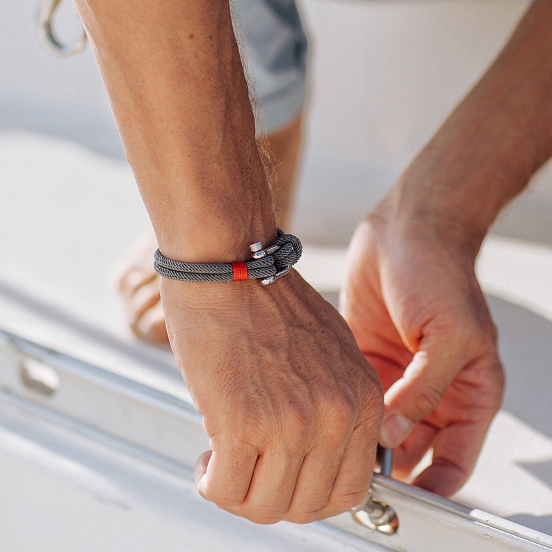 May include: A gray rope bracelet with a red accent and a silver anchor clasp.