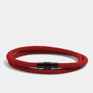 Puede incluir: Un brazalete de cuerda roja con un cierre de metal negro. El cierre tiene la palabra "Elevate" grabada.