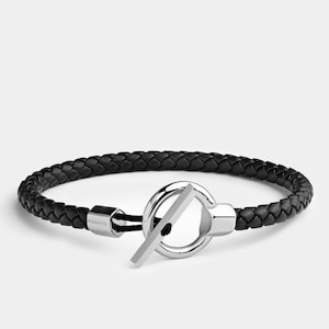 May include: A black leather bracelet with a silver clasp. The clasp is a ring with a bar across it. The bracelet is braided and has the word "Elegatto" engraved on the clasp.