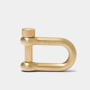 Könnte beinhalten: Ein goldfarbener Metallverschluss mit einer gerändelten Schraube und dem Wort "ELEGATTO" auf der Seite eingraviert.