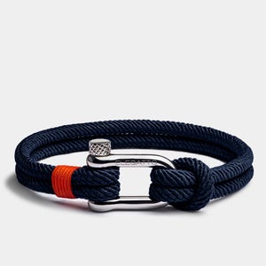 May include: Navy blue rope bracelet with a silver clasp. The bracelet features a bright orange accent and is knotted at the end. The word "ELEGATTO" is engraved on the clasp.