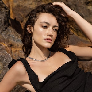 May include: A woman with curly brown hair wearing a black sleeveless top with a cowl neck. She is wearing a silver chain necklace.