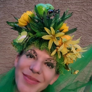 May include: A woman wearing a green and yellow floral crown with a green beetle on top. The crown is made of leaves, flowers, and netting. The woman is wearing green eye shadow and has a smile on her face.