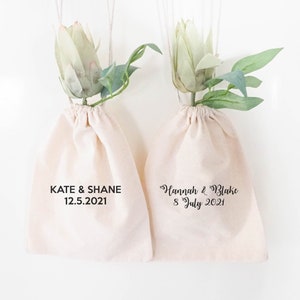 May include: Two white cotton drawstring bags with green floral accents. The bags are personalized with the names of couples and their wedding dates. The text on the bags reads "Kate & Shane 12.5.2021" and "Hannah & Blake 8 July 2021".