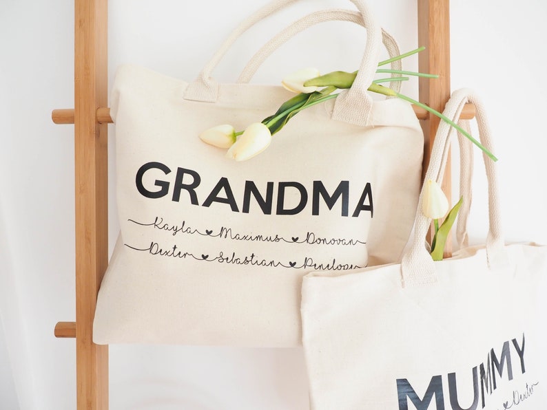 May include: Two white canvas tote bags with black handles. The larger bag says "GRANDMA" in bold black letters with the names "Kaylar, Maximus, Donovan, Dexter, Sebastian, Penelope" written in cursive underneath. The smaller bag says "MUMMY" in bold black letters with the name "Dexter" written in cursive underneath.