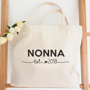 May include: A white canvas tote bag with black text that reads "NONNA est 2018" with a heart symbol between "est" and "2018".