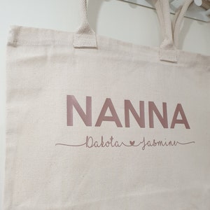 May include: A white canvas tote bag with the word "NANNA" printed in rose gold. Below the word "NANNA" is the text "Dakota &hearts; Jasmin" in rose gold.