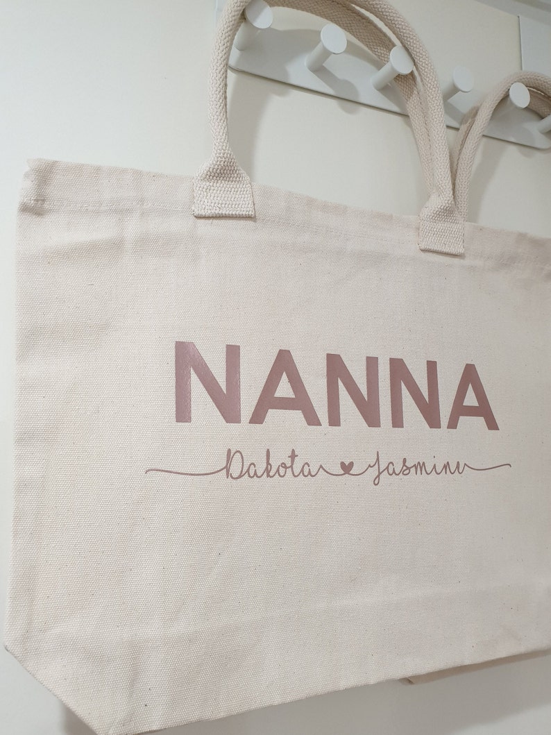 May include: A white canvas tote bag with the word "NANNA" printed in rose gold. Below the word "NANNA" is the text "Dakota &hearts; Jasmin" in rose gold.