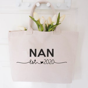 May include: A white canvas tote bag with black text that reads "NAN est 2020" with a heart symbol between "est" and "2020". The bag is hanging on a white door with a bouquet of white tulips inside.