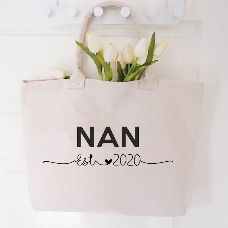 May include: A white canvas tote bag with black text that reads "NAN est 2020" with a heart symbol between "est" and "2020". The bag is hanging on a white door with a bouquet of white tulips inside.