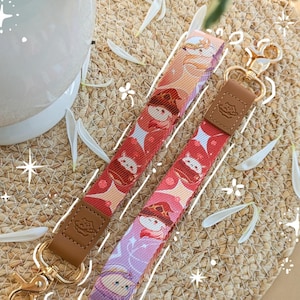 Puede incluir: Dos llaveros con patrones coloridos y personajes de dibujos animados. Tienen detalles de cuero marrón y cierres dorados. El fondo es una estera tejida con pétalos blancos dispersos. El sitio web "kuurimu.com" es visible en la parte inferior.