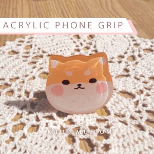 May include: A clear acrylic phone grip shaped like a cartoon Shiba Inu dog. The dog is orange and white with black eyes and a pink nose.