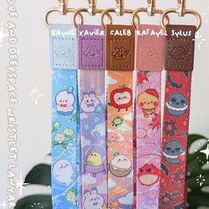 May include: Five fabric wristlet lanyards with gold-tone metal clasps and leather-like tabs embossed with a planet icon. Each strap features unique cartoon character prints and the names Zayne, Xavier, Caleb, Rafayel, and Sylus. Text reads LOVE AND DEEPSPACE WRISTLET LANYARD and KUURIMU.COM.