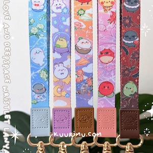 May include: Five fabric wristlet lanyards featuring colorful cartoon character prints, including space-themed and floral motifs. Each strap has a faux leather tab embossed with a planet logo and a gold-toned metal swivel lobster clasp at the base.