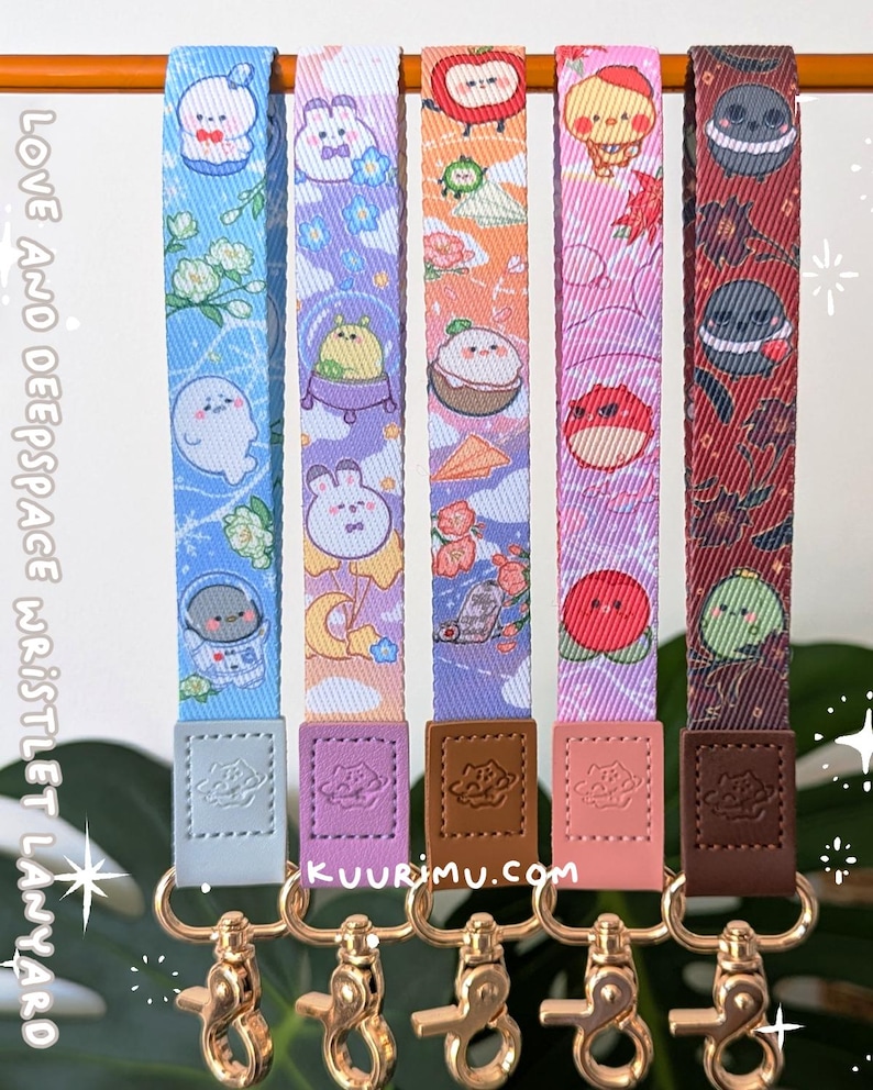 May include: Five fabric wristlet lanyards featuring colorful cartoon character prints, including space-themed and floral motifs. Each strap has a faux leather tab embossed with a planet logo and a gold-toned metal swivel lobster clasp at the base.