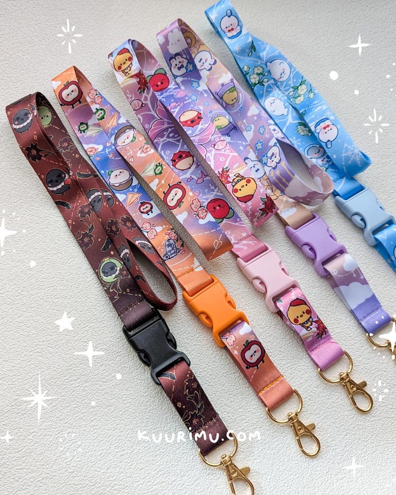 May include: Five fabric lanyards featuring colorful cartoon character prints and plastic side-release buckles. The lanyards come in brown, orange, pink, purple, and blue, each with a gold-toned metal swivel lobster clasp and the text kuurimu.com.