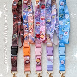 May include: Five fabric lanyards with plastic side-release buckles and gold-tone metal swivel hooks. Each features unique cartoon character prints in brown, orange, pink, purple, and blue. Text on the left reads LOVE, AND DEEPSPACE LANYARD.