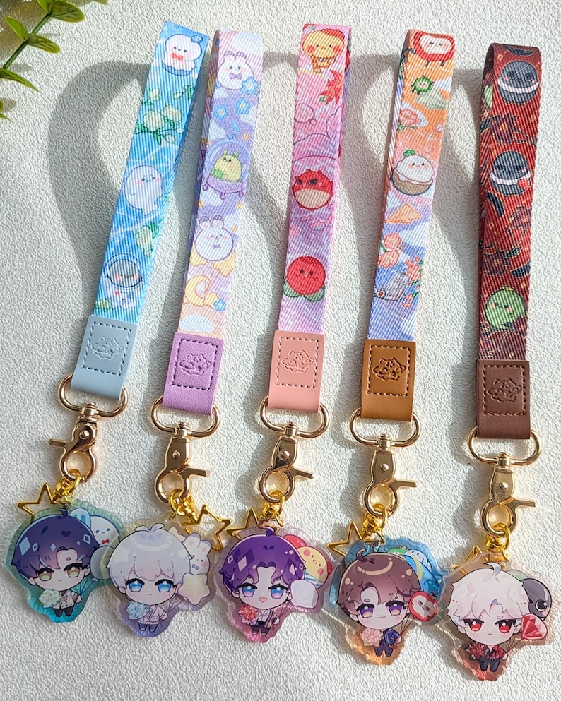 May include: Five fabric wristlet lanyards with gold-tone metal lobster clasps and faux leather tabs. Each lanyard features a unique colorful pattern with cartoon characters and is attached to a clear acrylic charm depicting an anime-style character.