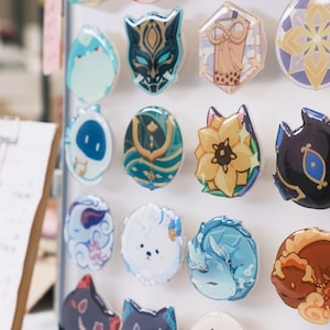 May include: A collection of colorful, round pins with various designs, including a cat wearing a mask, a flower, a cloud, and a dog. The pins are displayed on a white background.