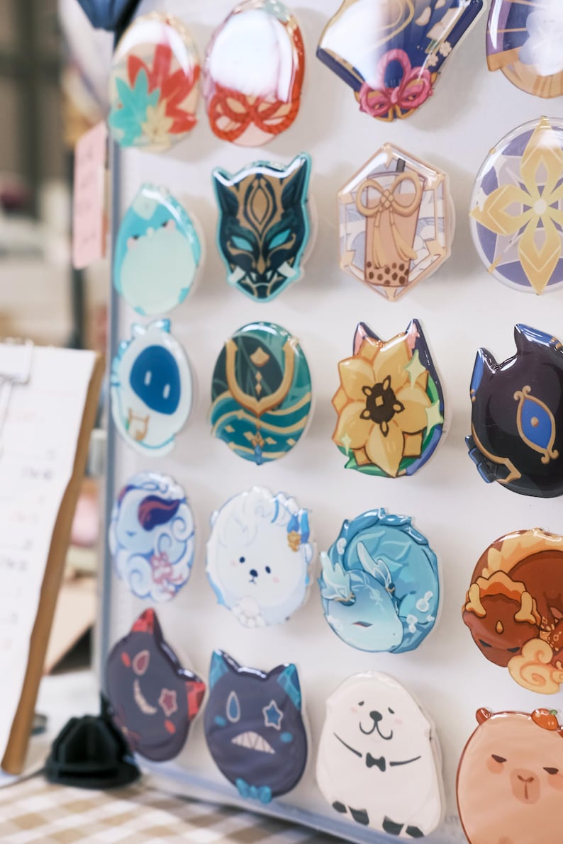 May include: A collection of colorful, round pins with various designs, including a cat wearing a mask, a flower, a cloud, and a dog. The pins are displayed on a white background.