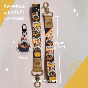 Pode incluir: Um cordão de pulso e um porta-chaves a condizer com um padrão de coelho de desenho animado. O cordão, com cerca de 18 cm de comprimento, tem ferragens douradas e um berloque de coelho a condizer. O texto "BANGBOO WRISTLET LANYARD" é visível.
