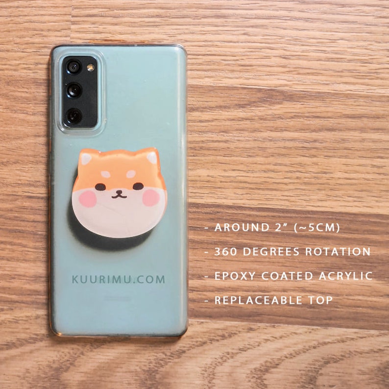 May include: A clear phone case with a Shiba Inu phone grip. The grip is approximately 2 inches in diameter and rotates 360 degrees. It is made of epoxy-coated acrylic and has a replaceable top. The phone case has the text "KUURIMU.COM" printed on the back.