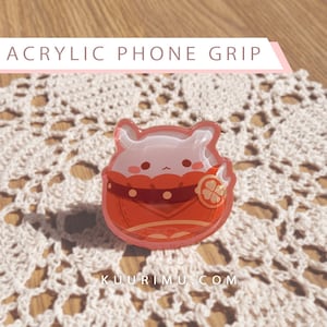 May include: A red and white acrylic phone grip with a cartoon character design. The character has a white body, red clothing, and a small orange fruit on its chest. The text "KUURIMU.COM" is visible at the bottom of the image.