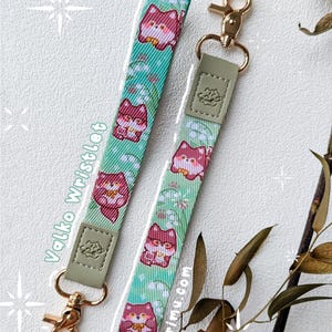 May include: Two wristlet lanyards featuring a repeating pattern of red fox characters and white lily of the valley flowers on a light green background. Each strap has a sage green faux leather tab with an embossed logo and a gold metal swivel clasp.