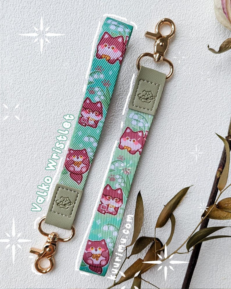 May include: Two wristlet lanyards featuring a repeating pattern of red fox characters and white lily of the valley flowers on a light green background. Each strap has a sage green faux leather tab with an embossed logo and a gold metal swivel clasp.