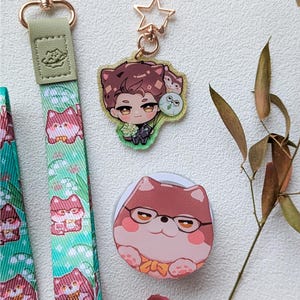 May include: A green fabric lanyard with a repeating pattern of cartoon animals, a gold-tone lobster clasp, a star-shaped keychain featuring an illustrated character with brown hair, and a circular accessory with a cartoon animal wearing glasses.