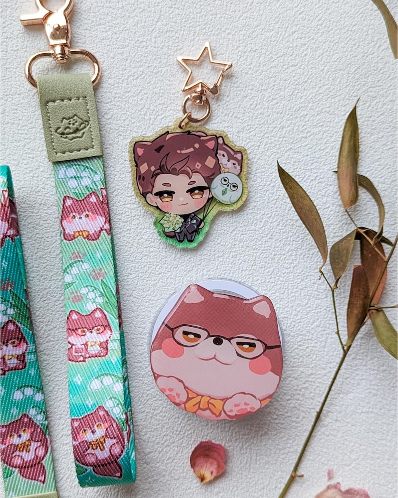 May include: A green fabric lanyard with a repeating pattern of cartoon animals, a gold-tone lobster clasp, a star-shaped keychain featuring an illustrated character with brown hair, and a circular accessory with a cartoon animal wearing glasses.