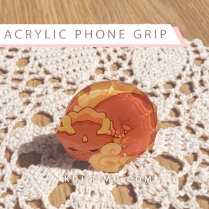 May include: A red and yellow acrylic phone grip with a cartoon dragon design. The grip is shaped like a circle and has a glossy finish.