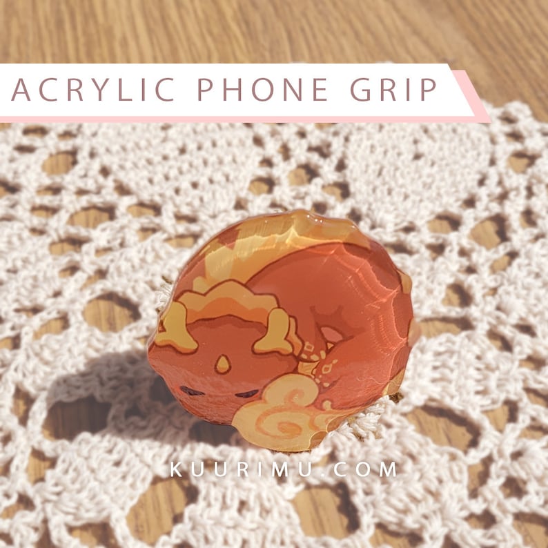 May include: A red and yellow acrylic phone grip with a cartoon dragon design. The grip is shaped like a circle and has a glossy finish.