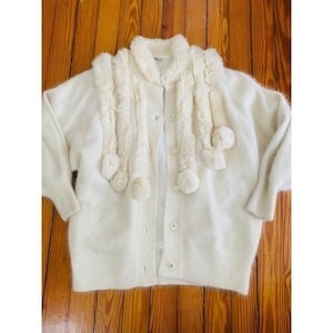 May include: Cream-colored cardigan with a matching faux fur scarf. The cardigan has a button-up front and long sleeves. The scarf features long, fluffy strands and pom-poms at the ends. The garment is displayed on a wooden surface.